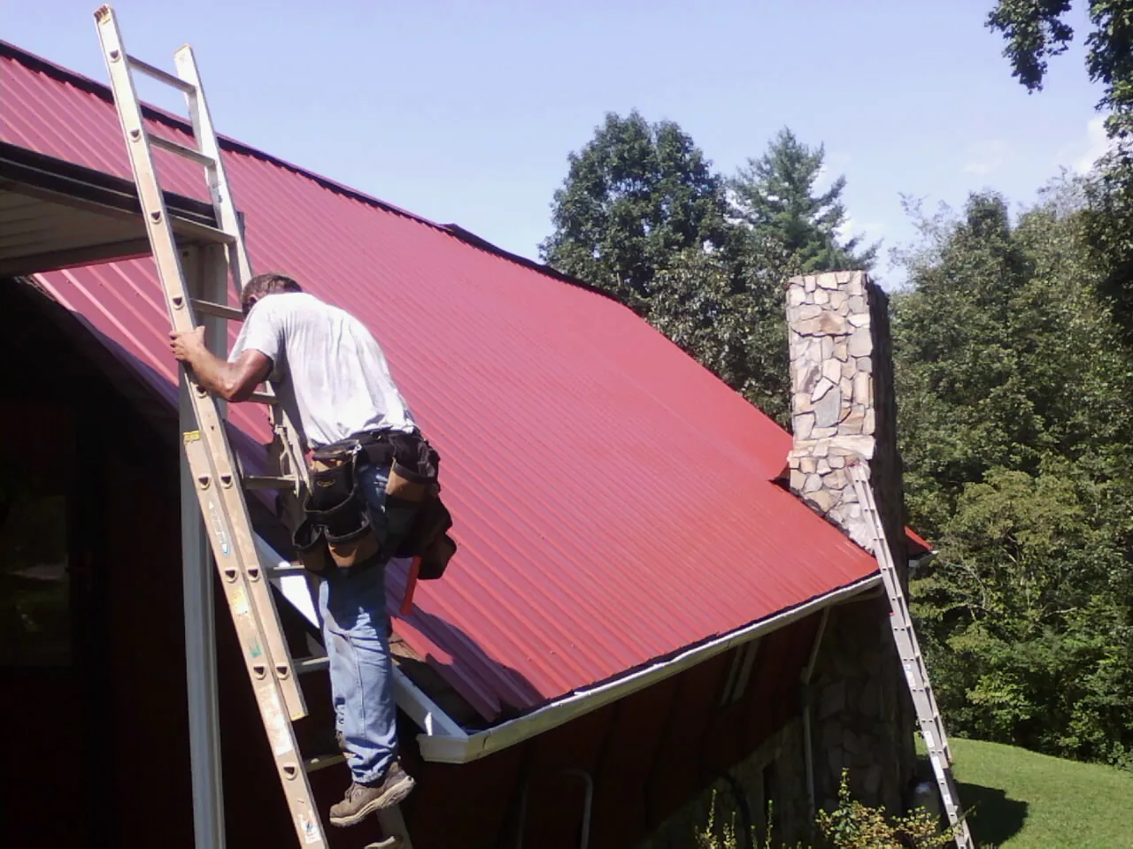 Roofer on a ladder beside a red metal roof and stone chimney — Soffit & Fascia Repair by Roofing Brothers in Canaan